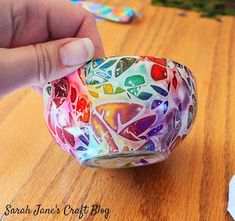 a hand holding a colorful glass bowl on top of a wooden table