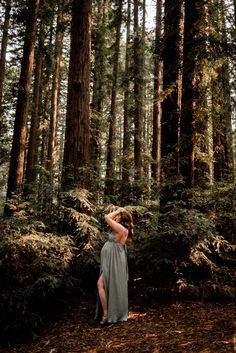 a woman standing in the middle of a forest wearing a gray dress and holding her hair back