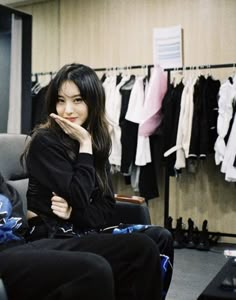 a woman sitting on a chair in front of a rack of clothes with her chin resting on her hand