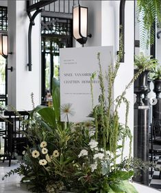 an arrangement of flowers and greenery is displayed in front of a sign that reads