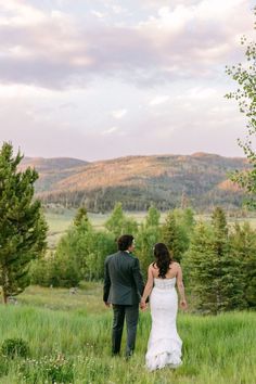 a bride and groom holding hands walking through tall grass with mountains in the back ground