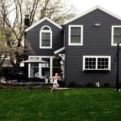 a woman is walking in front of a gray house with white trim on the windows