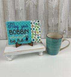 there is a coffee mug next to a card on a stand with the words may your bobbin always be full