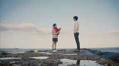 two people standing on rocks near the ocean and one person is holding something in his hand