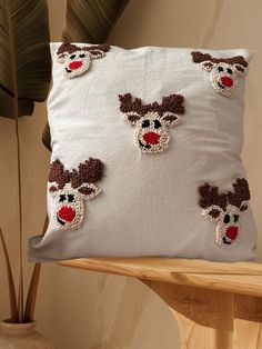 a white pillow with reindeer head appliqued on the front and sides, sitting on a wooden table next to a potted plant