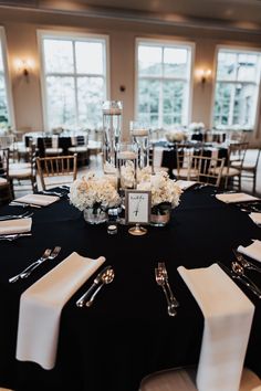 a black table with white flowers and silverware on it in front of two large windows