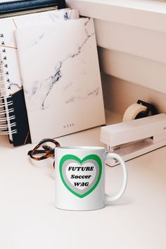 a white coffee mug sitting on top of a desk next to a binder and eyeglasses