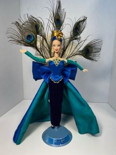 a doll with peacock feathers on it's head and blue dress, standing in front of a white background