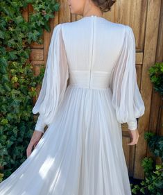 a woman wearing a white dress standing in front of a wooden wall with ivy growing on it