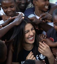 a woman is surrounded by children in a group smiling and touching her hair with both hands