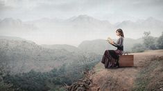 a woman sitting on top of a cliff reading a book with mountains in the background