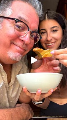 a man and woman are eating food from a white bowl while smiling at the camera