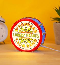 a clock sitting on top of a wooden table next to a potted plant and a yellow wall