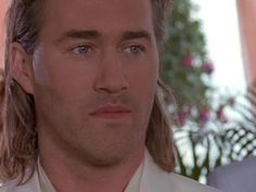 a man with long hair wearing a white shirt and tie in front of a potted plant