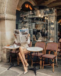 a woman sitting at an outdoor cafe reading the paper while holding a cup of coffee