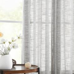 a white vase with flowers on a table in front of a window that has sheer curtains