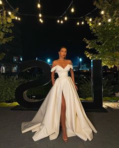 a woman standing in front of a sign wearing a long white dress with high slit