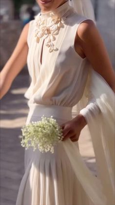 a woman wearing a white dress and holding a flower bouquet in her hand while walking down the street