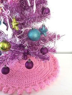 a pink crocheted christmas tree skirt with ornaments on the bottom and purple balls hanging from it