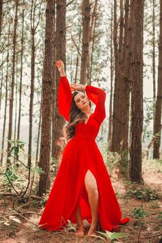 a woman in a red dress is standing in the woods with her arms up and hands behind her head