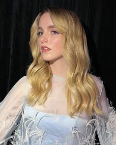 a woman with long blonde hair wearing a white dress and feathered sleeves, standing in front of a black backdrop