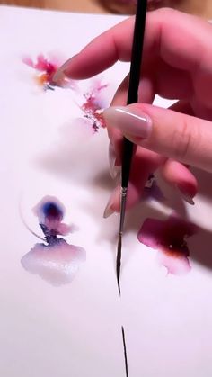 a woman's hand holding a pen and drawing flowers on paper with watercolors