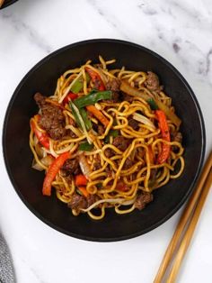 a black bowl filled with noodles and meat next to chopsticks on a white table