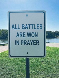 a white sign that says all battles are won in prayer on the grass next to water