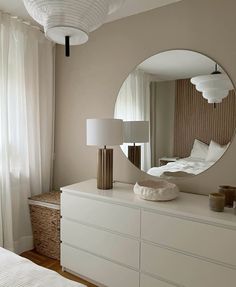 a white dresser topped with a round mirror next to a lamp and a large window