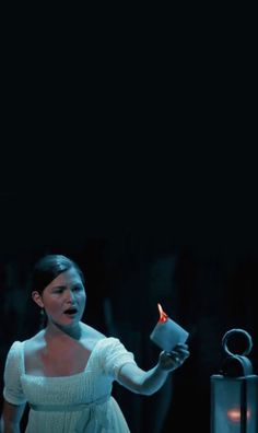 a woman in white dress holding a lit candle and looking at the ground with her mouth open