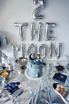 a table topped with a blue cake and lots of silver balloons that spell out the word's the moon