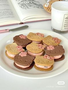 a white plate topped with cookies covered in pink and brown frosting next to an open book