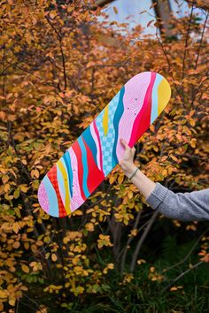 a woman holding up a colorful skateboard in front of some trees with yellow leaves