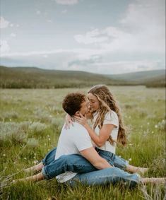 a man and woman sitting on the ground kissing in a field with mountains in the background