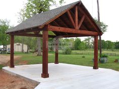 a wooden gazebo sitting on top of a cement slab next to a green field