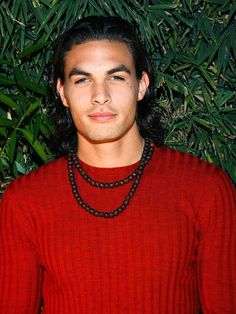 a man with long hair wearing a red sweater and beaded necklace standing in front of plants