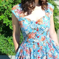 a woman wearing a blue floral dress standing in front of some bushes and flowers with her hands on her hips