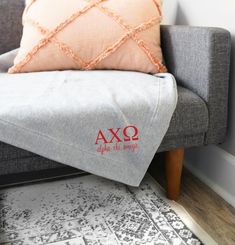 a grey couch with a red axo embroidered pillow on it's back next to a white rug