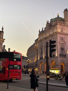 a red double decker bus driving down a street next to tall buildings and traffic lights