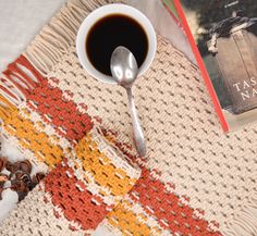 a cup of coffee next to a book and spoon on a knitted tablecloth