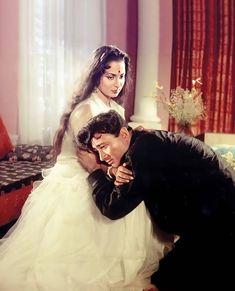 a man kneeling down next to a woman in a white dress on a bed with red curtains