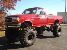 a red pick up truck with big tires parked in a parking lot next to a tree