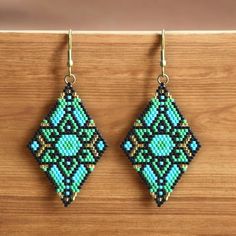 This pair of intricately handcrafted earrings features a beautiful geometric diamond design made from tiny, colorful beads. The pattern is carefully arranged with vibrant shades of green, blue, black, and accents of gold for a touch of elegance. These earrings are lightweight, versatile, and perfect for adding a bold, artistic statement to any outfit. Ideal for boho-chic, ethnic, or casual looks, they highlight exceptional craftsmanship and artistry. Crafted by Differently-abled Artist Geometric Diamond Design, Differently Abled, Geometric Diamond, Handcrafted Earrings, Diamond Design, Green And Blue, Shades Of Green