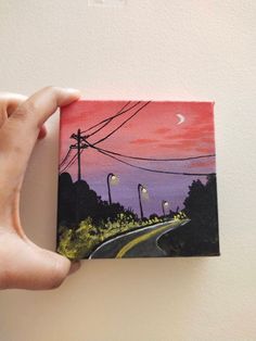 a person holding up a small painting on a wall with street lights in the background