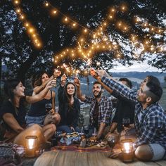 a group of people sitting around a picnic table with lights strung from the trees over them