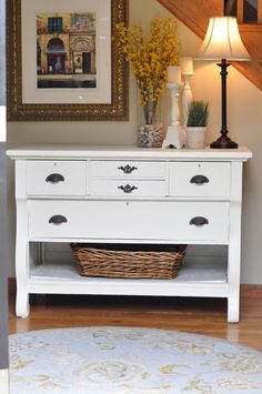 a white dresser with two drawers and baskets on it in front of a framed painting