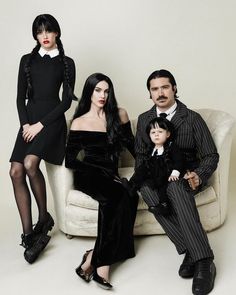 three people and a child sitting on a couch in front of a white wall wearing black clothing