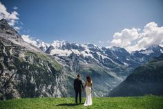 a bride and groom standing on top of a lush green hillside with mountains in the background