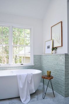 a white bath tub sitting under a window next to a wooden table with a gold cup on it