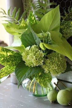 a glass vase filled with green flowers and greenery next to some pears on a table